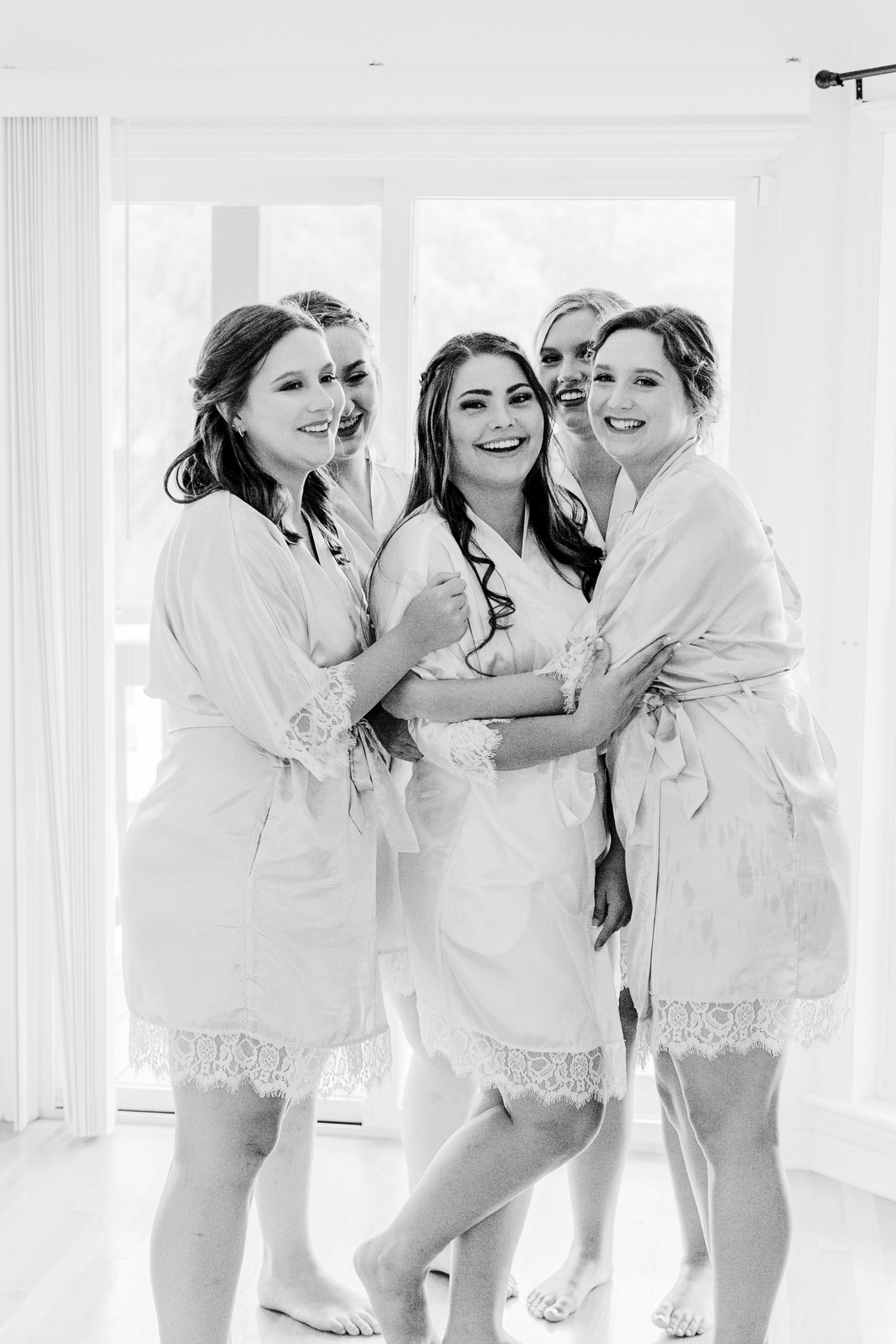 Black and white photo of a bride and four bridesmaids in matching lace robes smiling and embracing before the wedding.