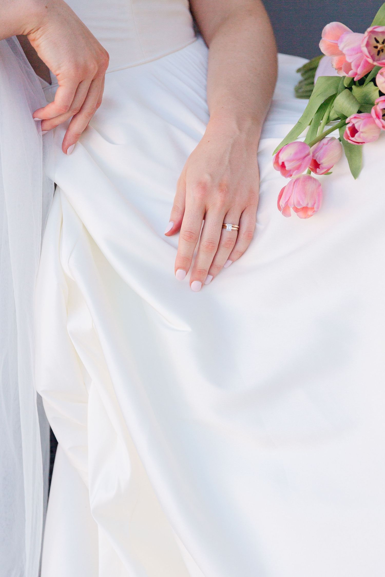 Elegant white wedding dress detail with pink tulips and diamond ring on fabric.