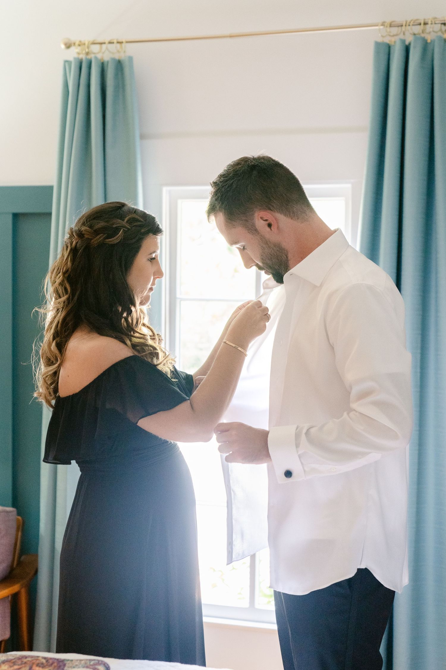 A couple in formal attire share a tender moment while getting ready in a room with turquoise curtains.