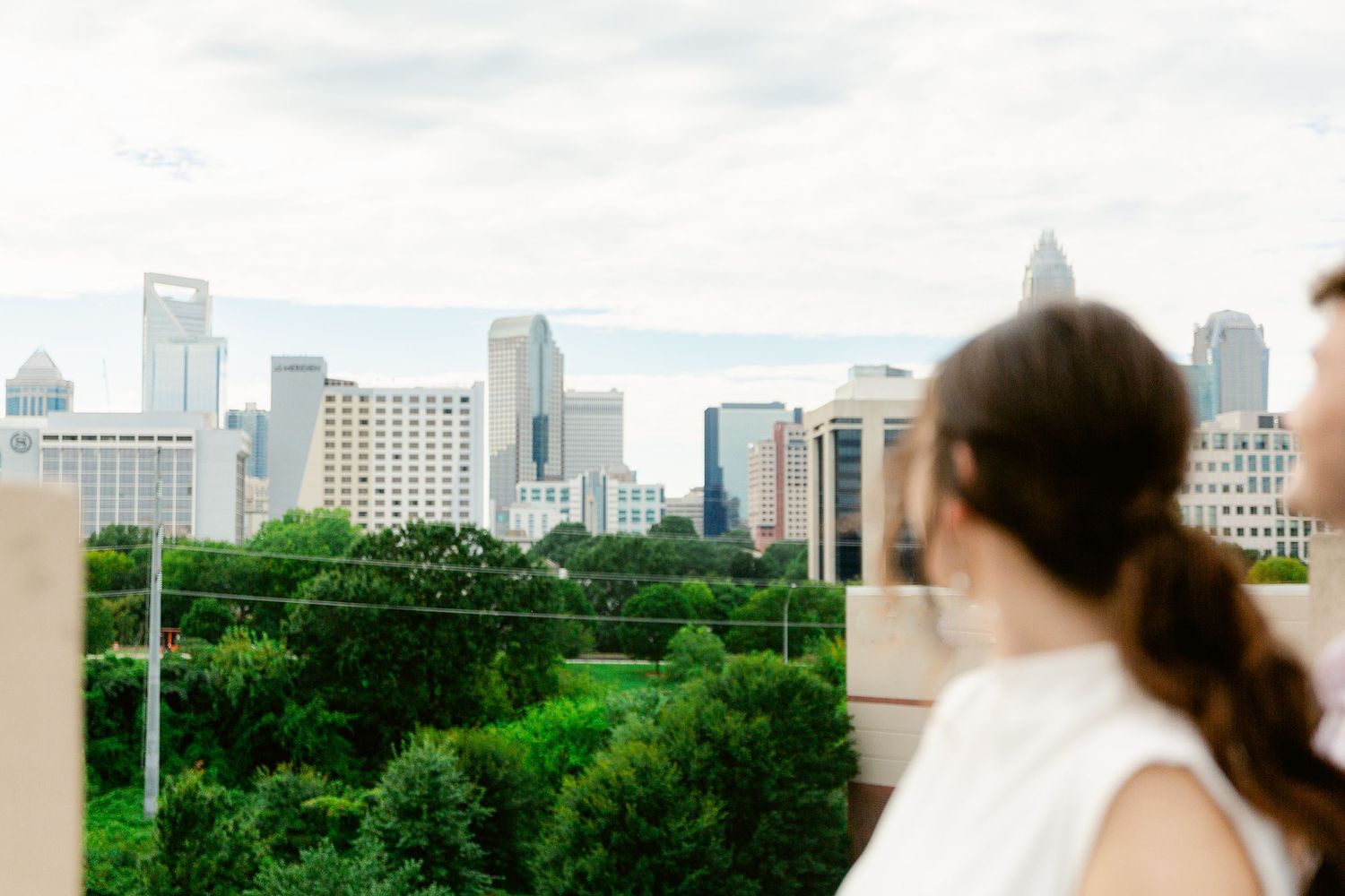 A person looks out at the Charlotte, North Carolina skyline with lush green trees in the foreground on a cloudy day.