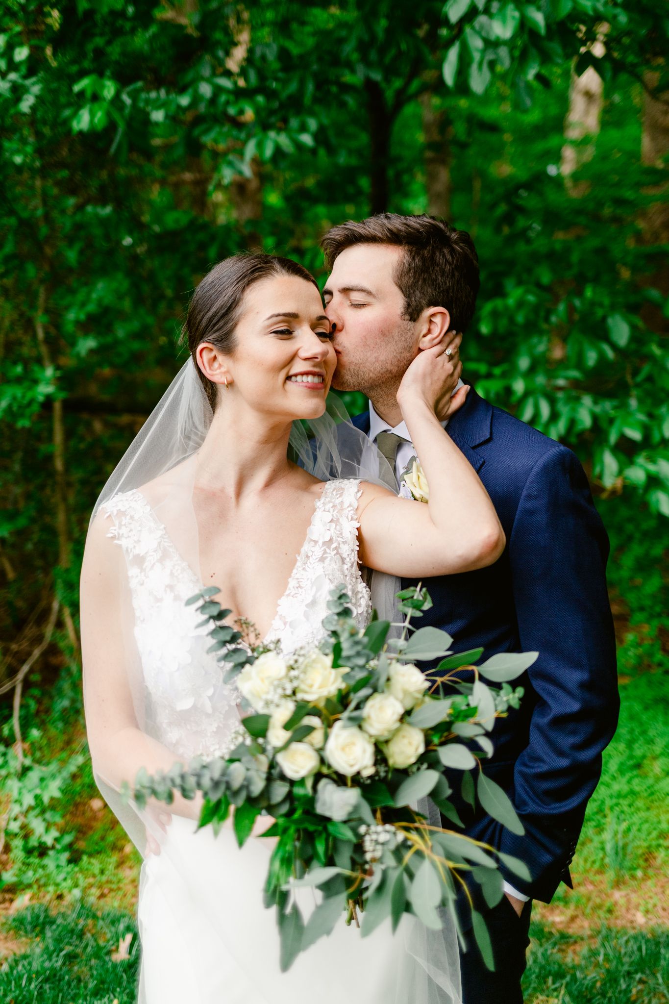 Romantic outdoor wedding photos showing couple embracing among lush greenery with white floral bouquet.