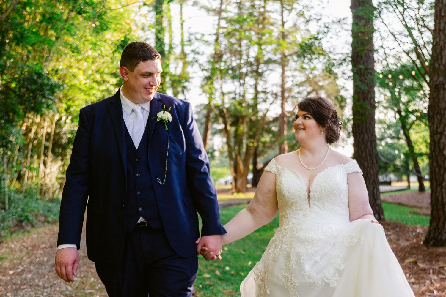 Newlyweds share a tender moment while walking through a tree-lined path in a park.
