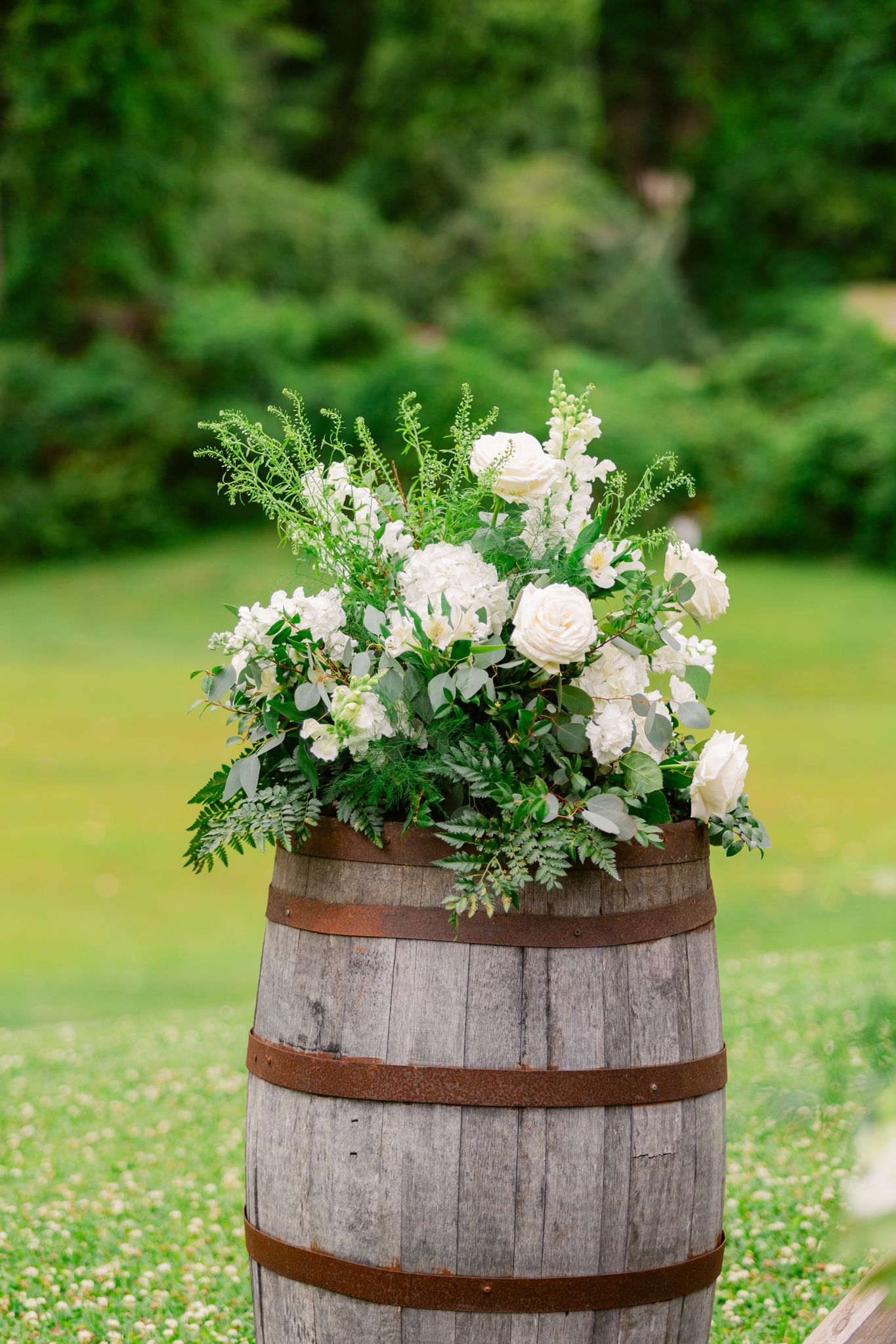 A rustic wooden wine barrel topped with an elegant floral arrangement of white roses and greenery in an outdoor garden setting.