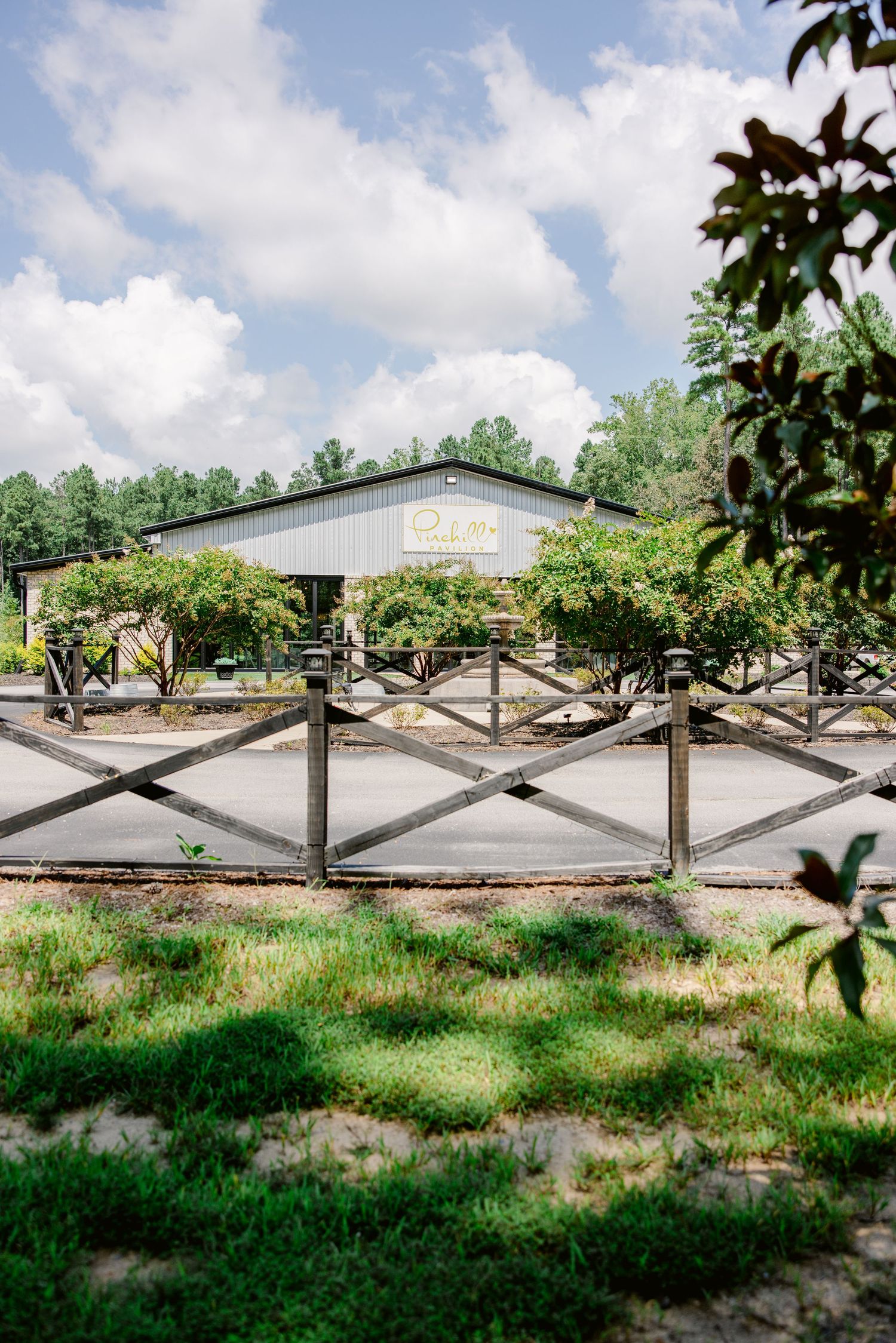 Wooden cross-beam fencing stretches along a grassy field with trees and a white building in the background.