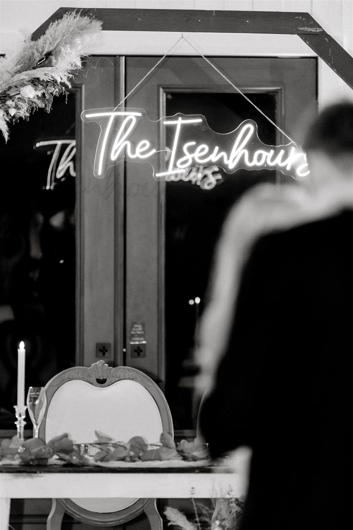 An illuminated neon sign reading 'The Isenhouse' with pampas grass decor in monochrome.