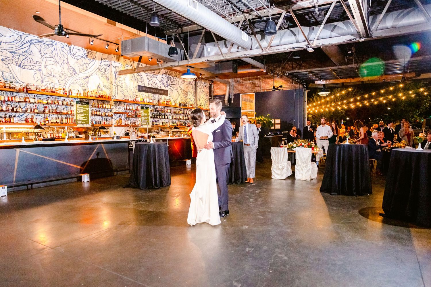 Couple dancing in an industrial-style venue with exposed ceiling and string lights during wedding reception.