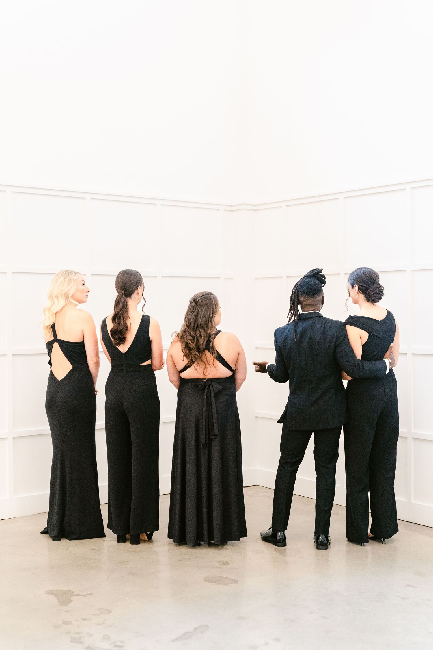 A group of people wearing black formal attire standing in a line against a white wall, photographed from behind.