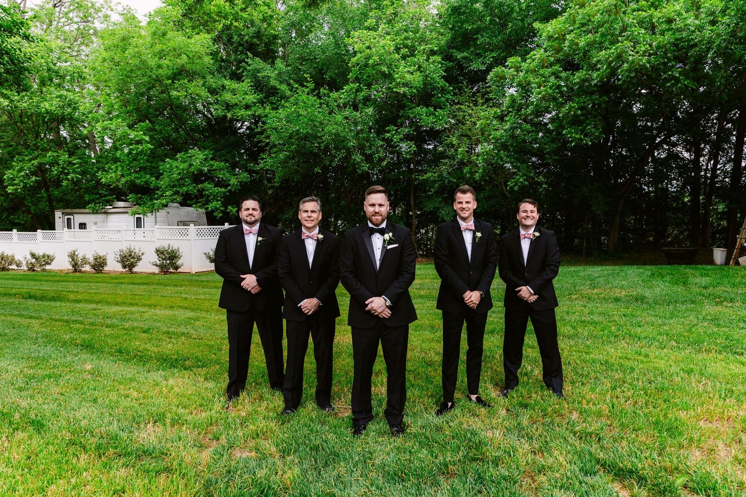 A group wearing formal black suits and ties stands in a lush green garden setting for a special occasion photo.