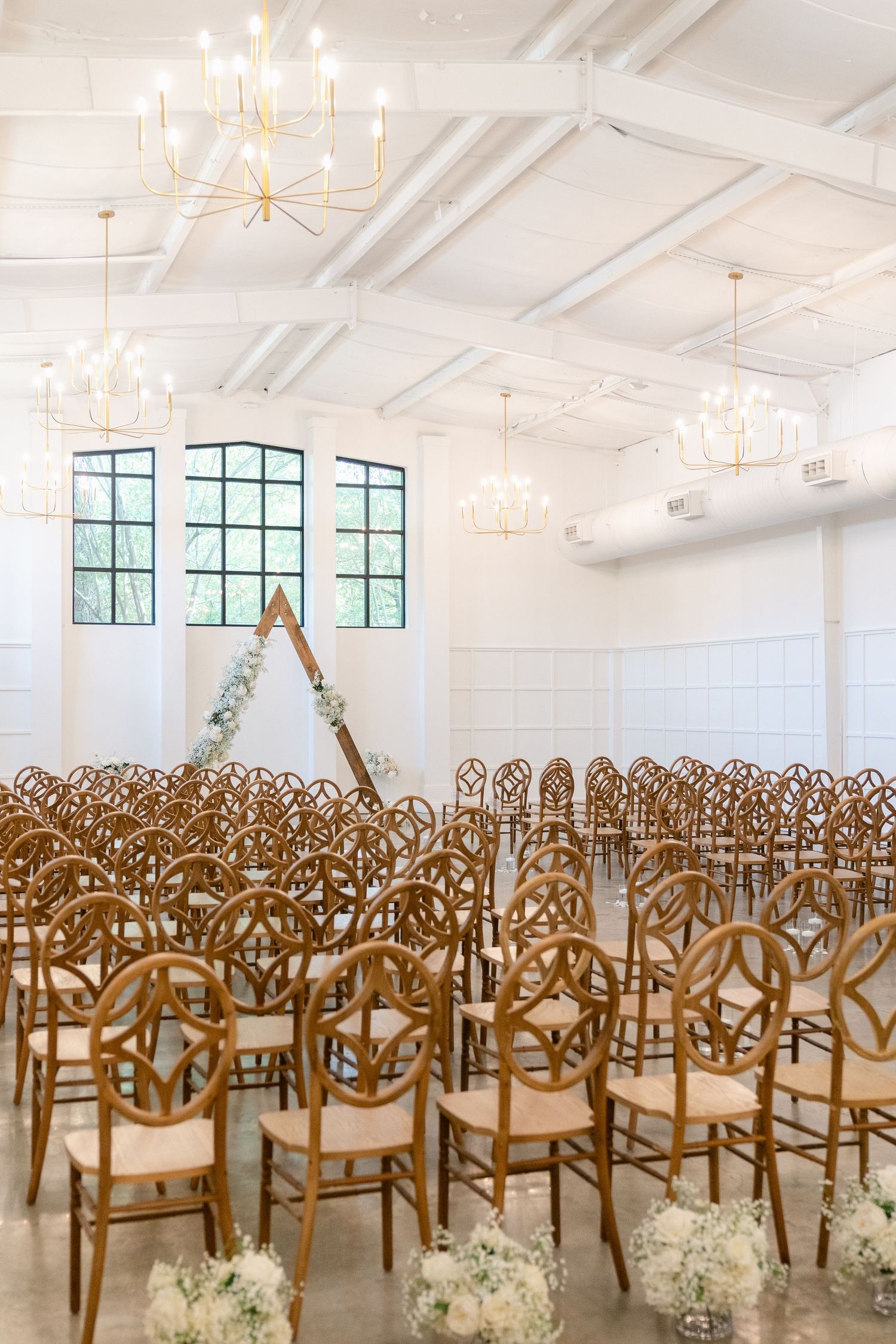 Elegant wedding ceremony setup with rows of ornate wooden chairs in a bright white room with vaulted ceilings and chandeliers.