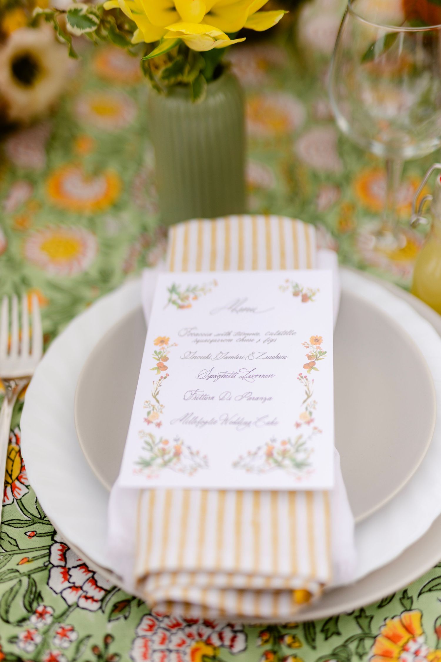 Elegant place settings with floral patterned cards and yellow flowers on a green tablecloth at a spring event.
