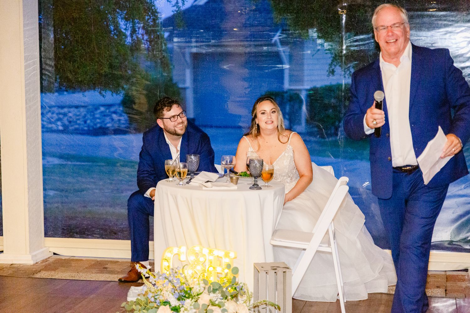 Wedding reception guests seated at outdoor tables with blue lighting and floral centerpieces at sunset.