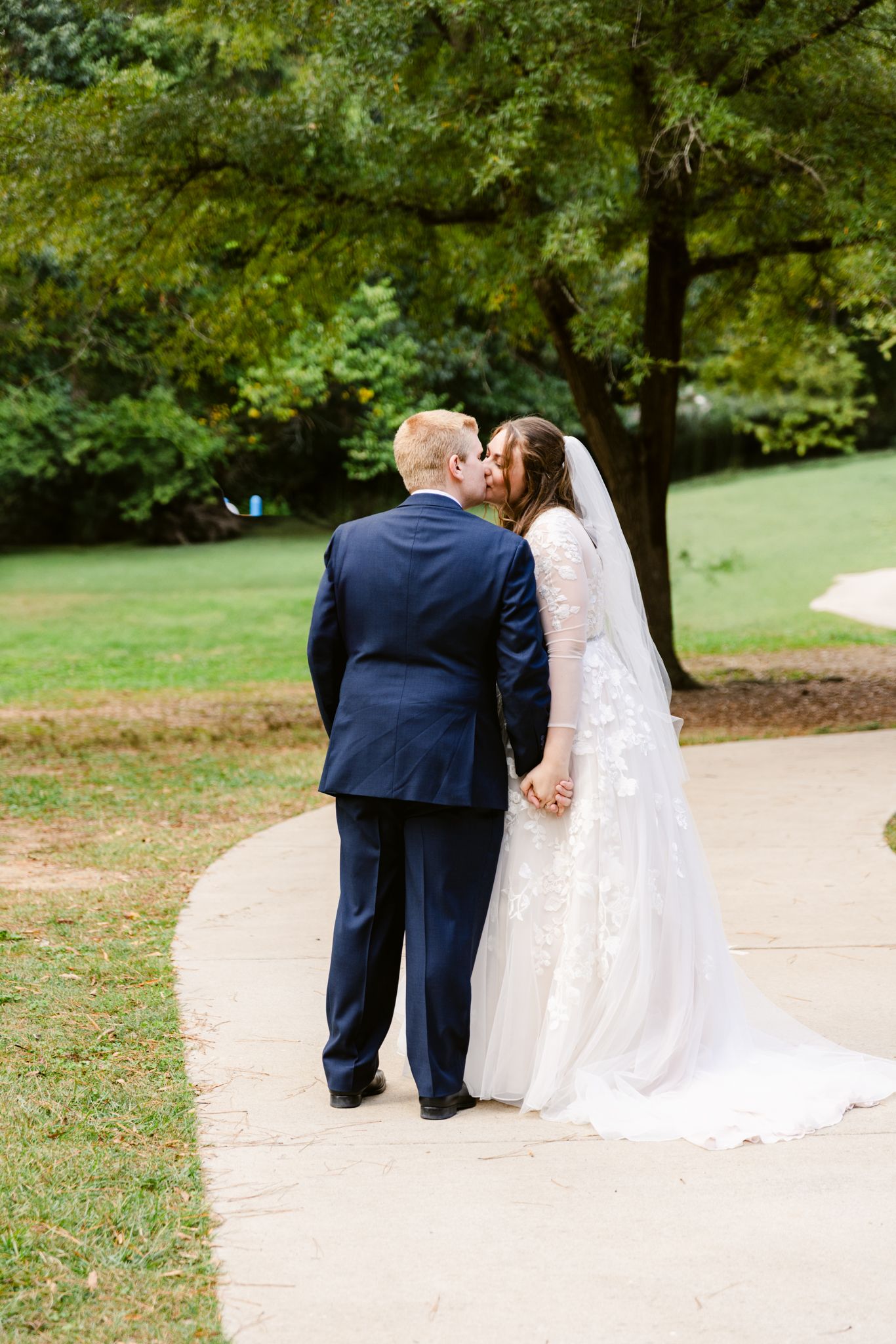 A couple in wedding attire shares a romantic moment on a winding path surrounded by lush green trees.