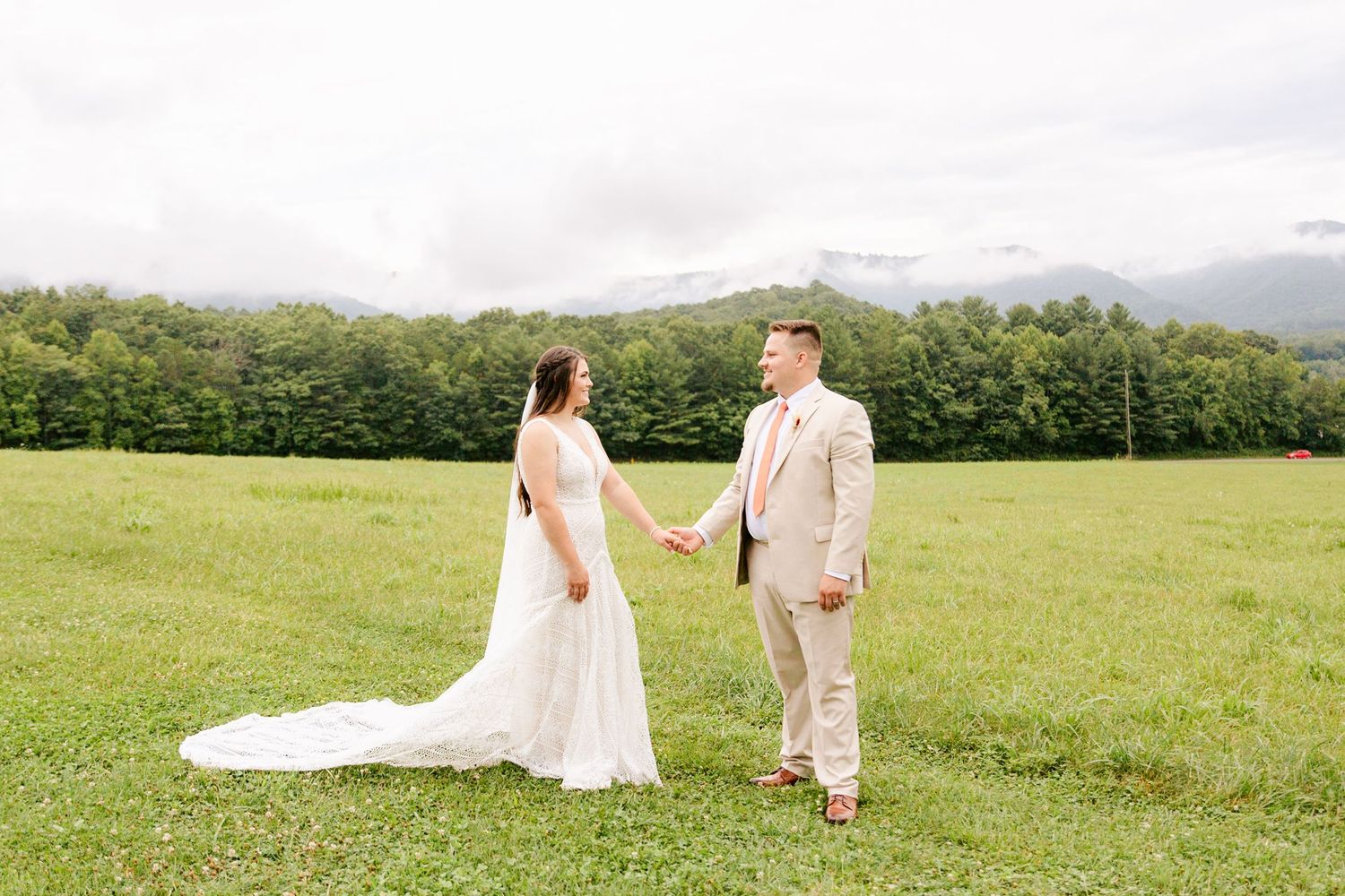 A romantic couple walks together across a scenic hilltop meadow with mountain views in the background.