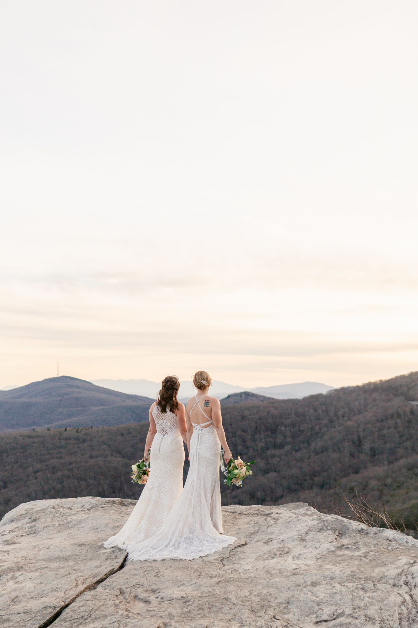 Two people in elegant white dresses stand together on a mountain overlook at sunset holding bouquets.