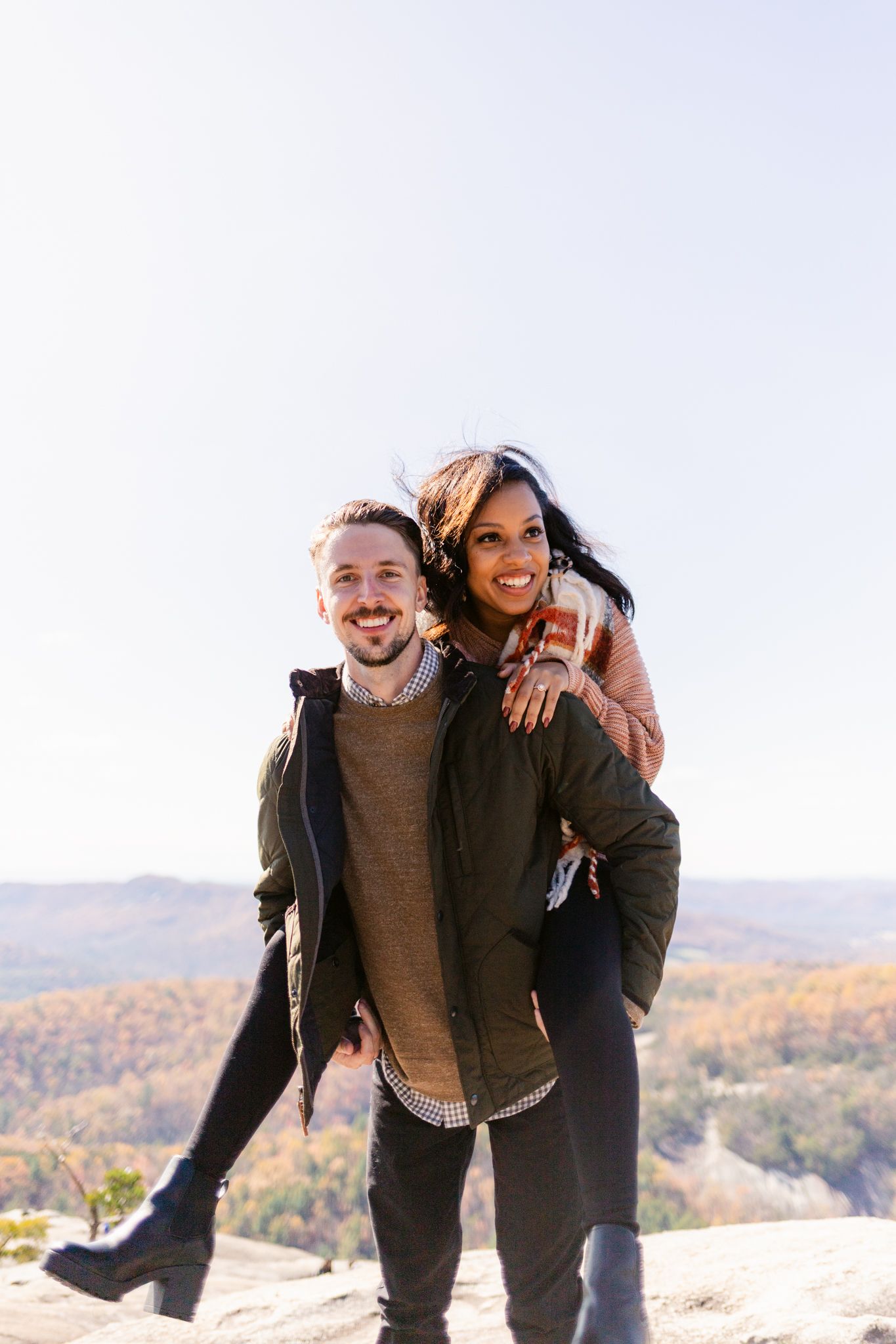 Couple enjoying a playful piggyback ride moment together on a mountain overlook with autumn foliage in background.