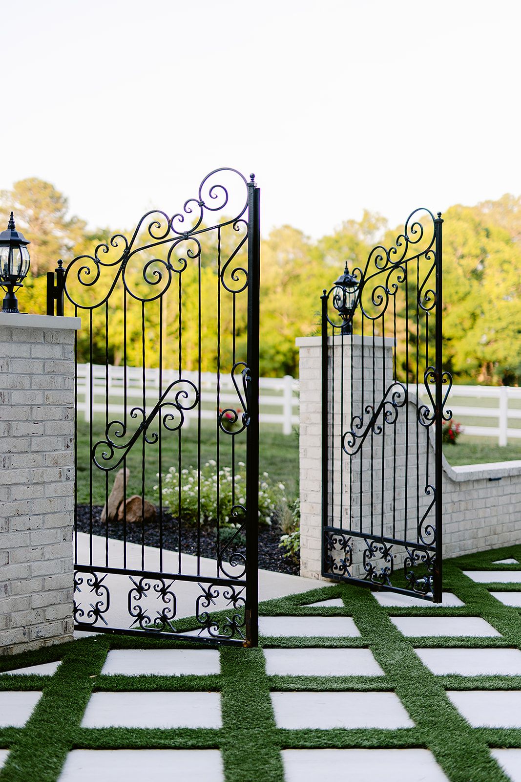Ornate black wrought iron garden gate with decorative scrollwork opens onto a geometric paver pathway with grass inlays.
