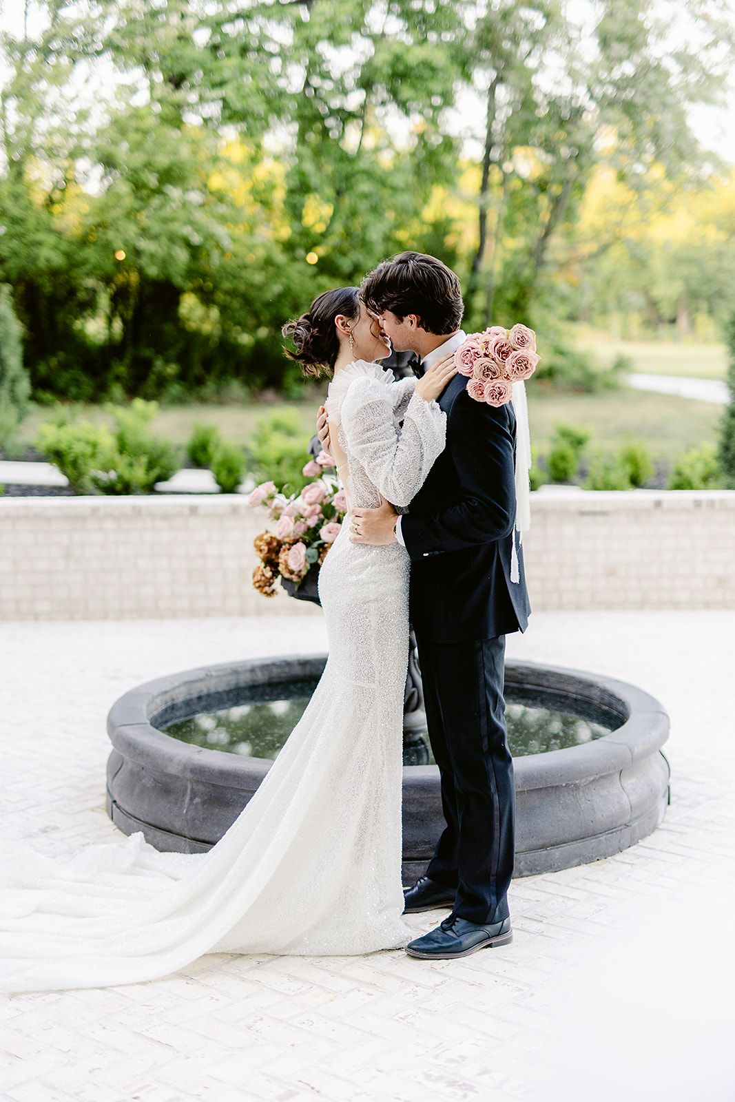 A romantic wedding couple embraces near a circular fountain, the bride in a flowing white gown holding pink roses.