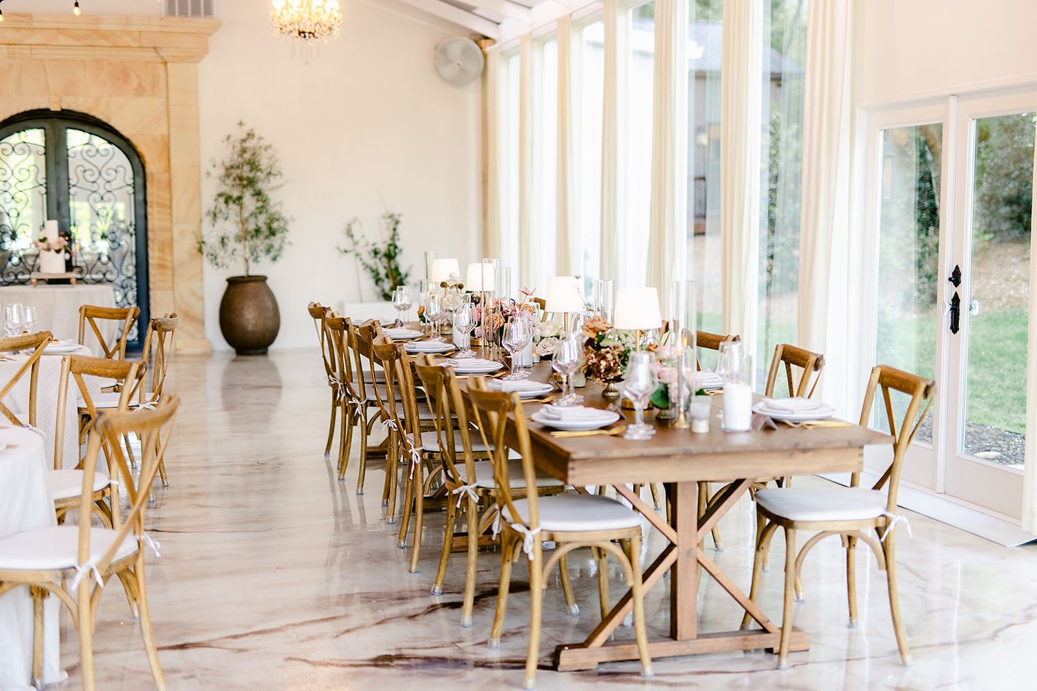 Bright white sunroom with wooden chairs and tables arranged for dining, featuring arched windows and elegant decor.