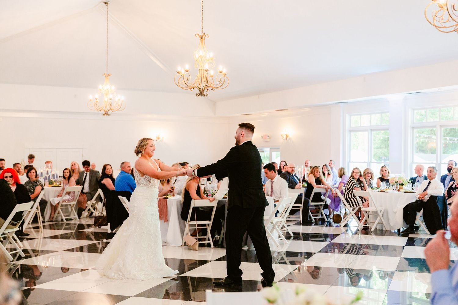 Wedding guests dance together on a white dance floor under elegant chandeliers in a reception tent.