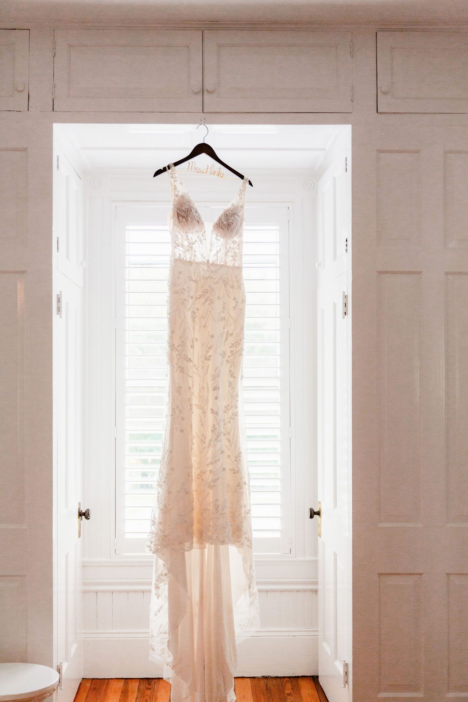 A delicate lace wedding dress hangs elegantly in front of white shuttered windows in natural light.