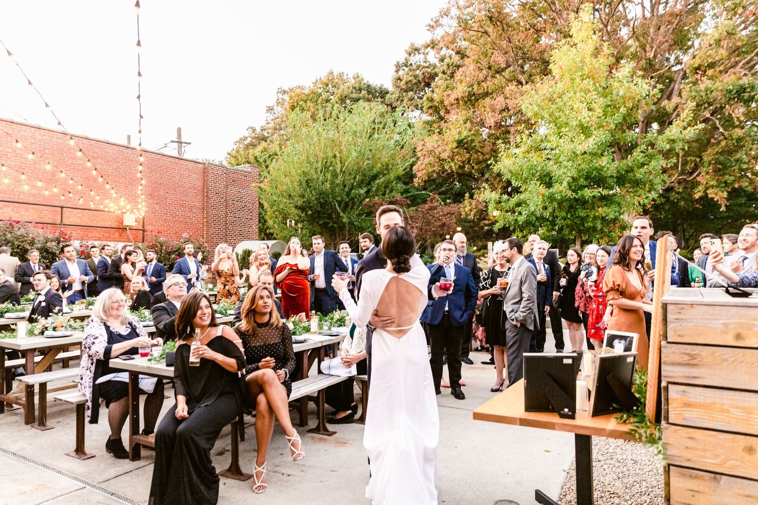 A crowd gathers outside a brick building with trees and landscaping during a daytime event.