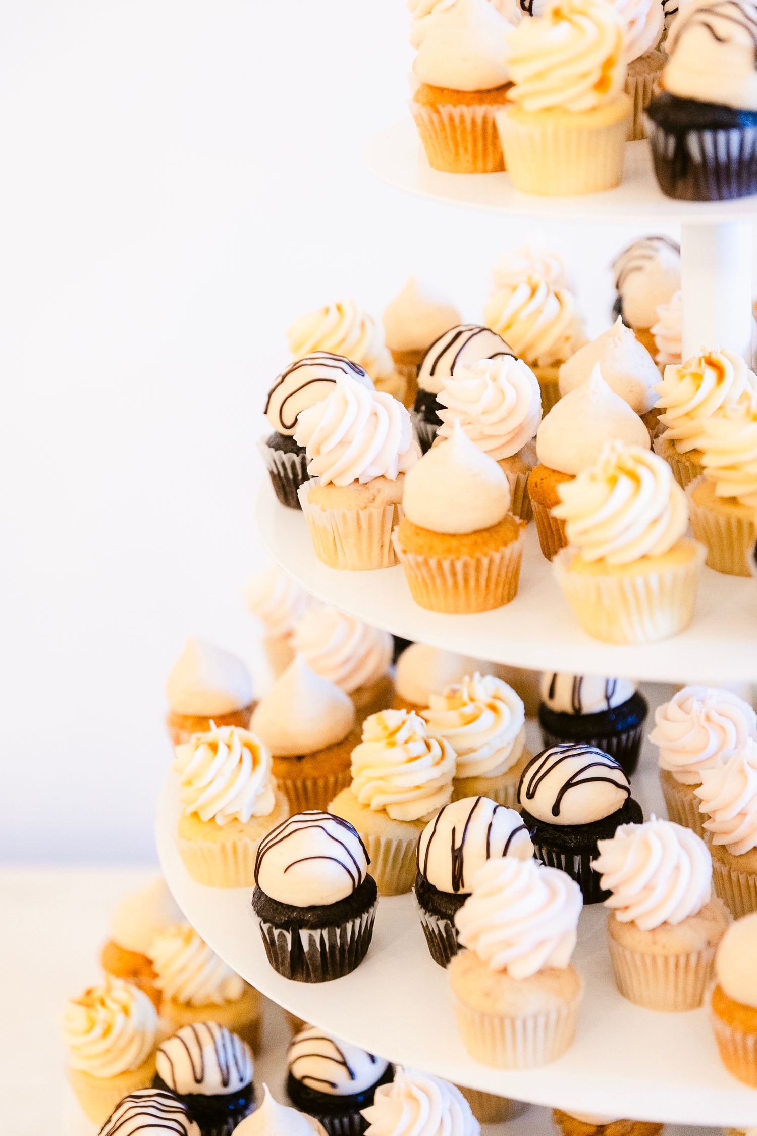 Elegant tiered display of vanilla and chocolate cupcakes with swirled frosting and drizzled toppings on a white stand.