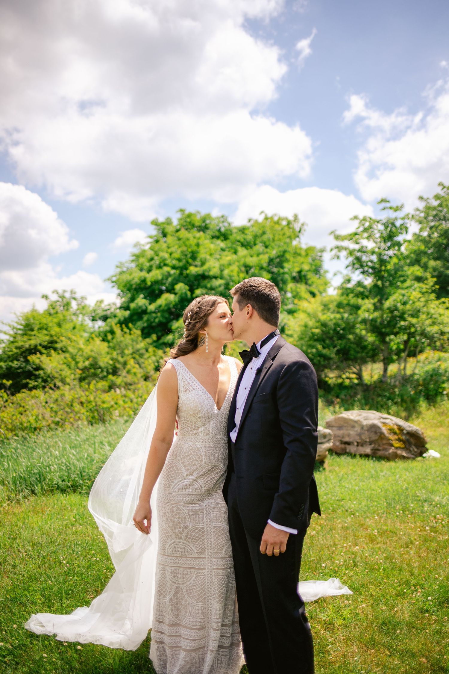 A romantic outdoor wedding portrait of a couple in formal attire with flowing veil against lush greenery.