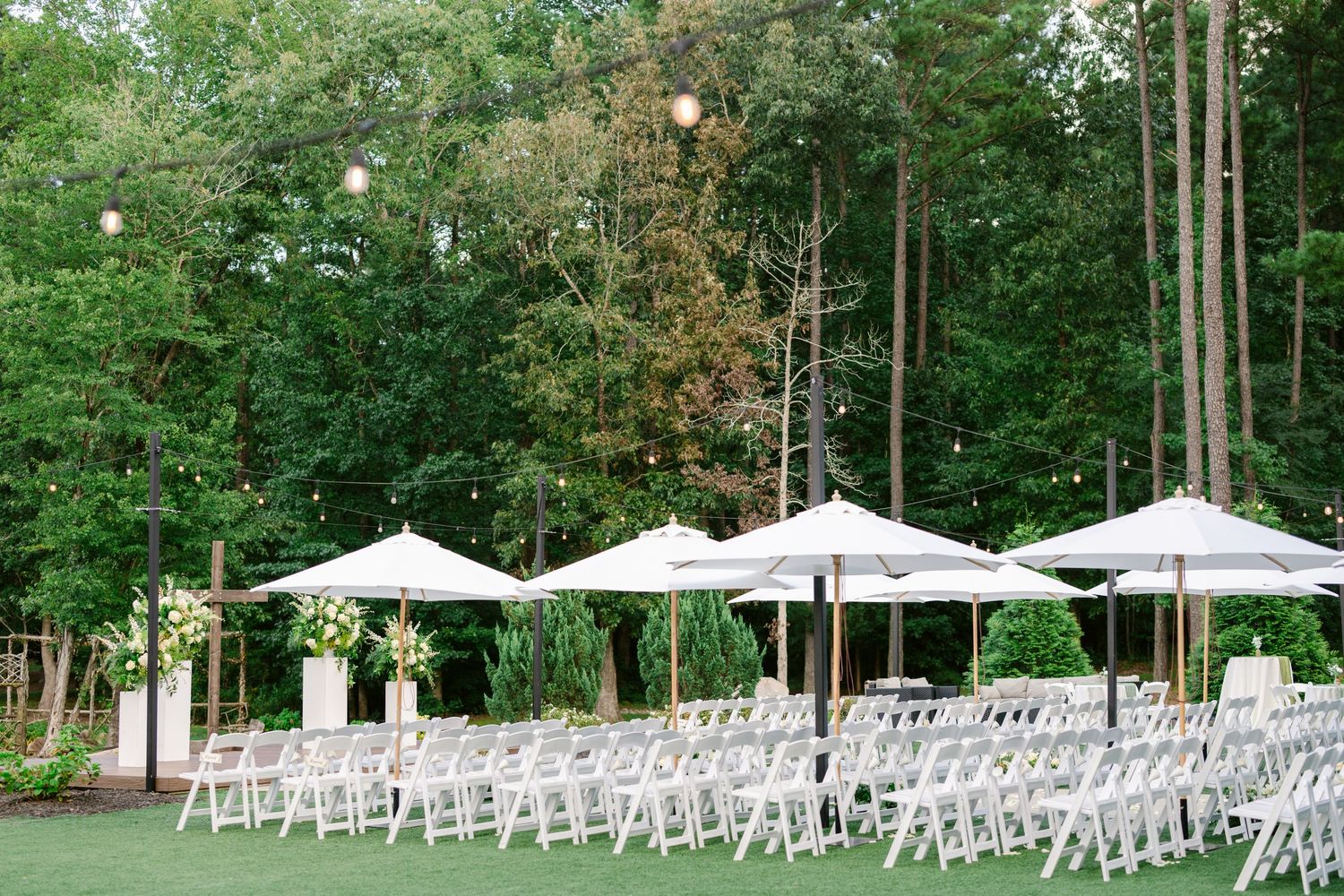 White chairs and umbrellas set up for an outdoor wedding ceremony in a wooded garden setting.