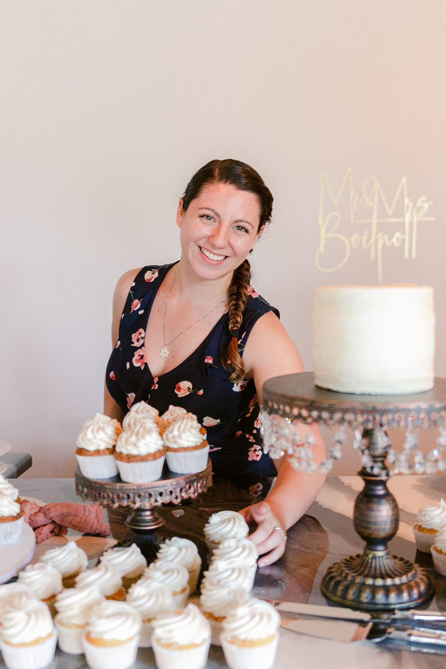 A series of cupcakes with white frosting are arranged next to an elegant white cake on a silver cake stand.