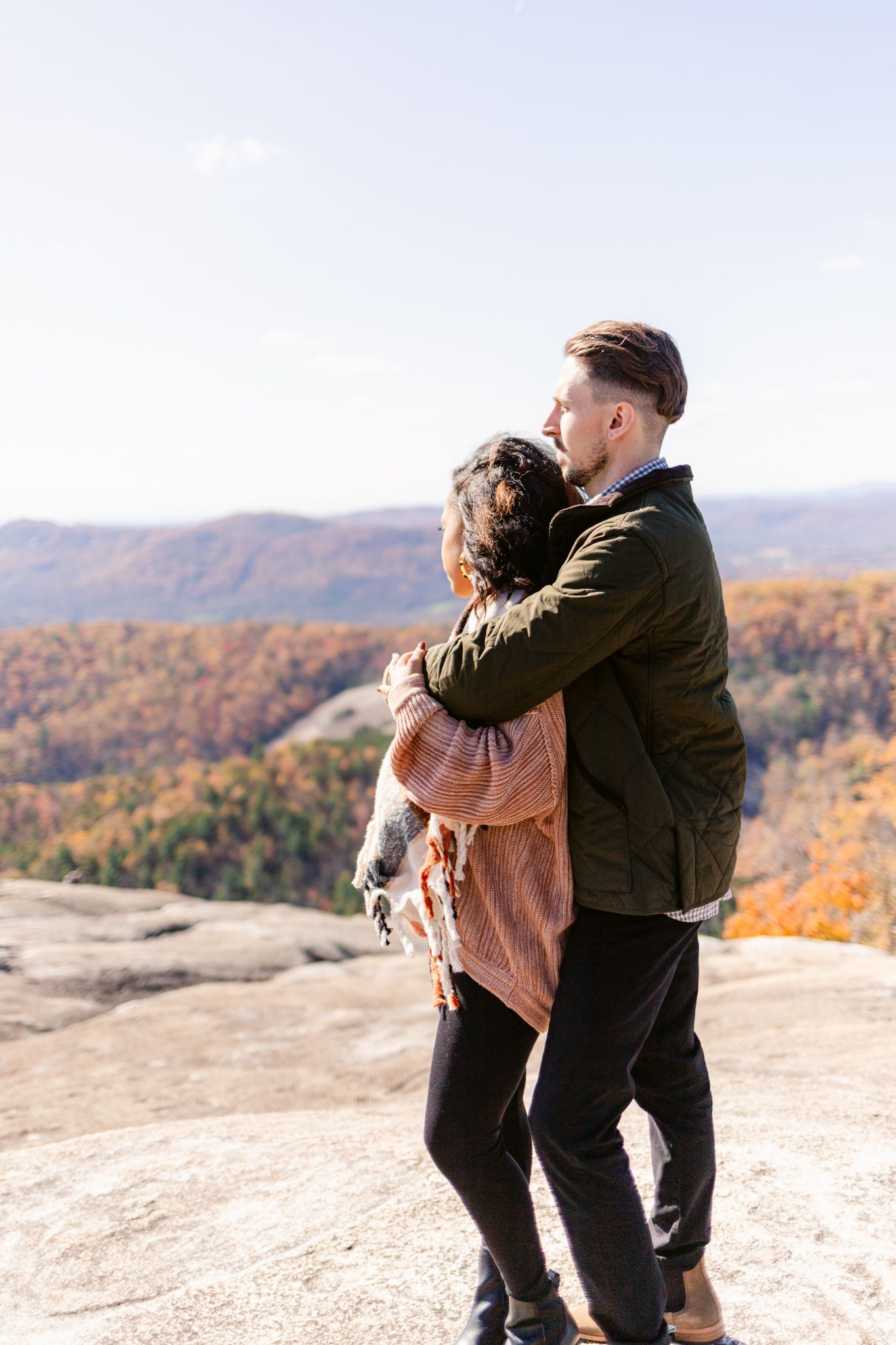 A couple embraces while standing on a rocky mountain overlook with autumn foliage in the background.