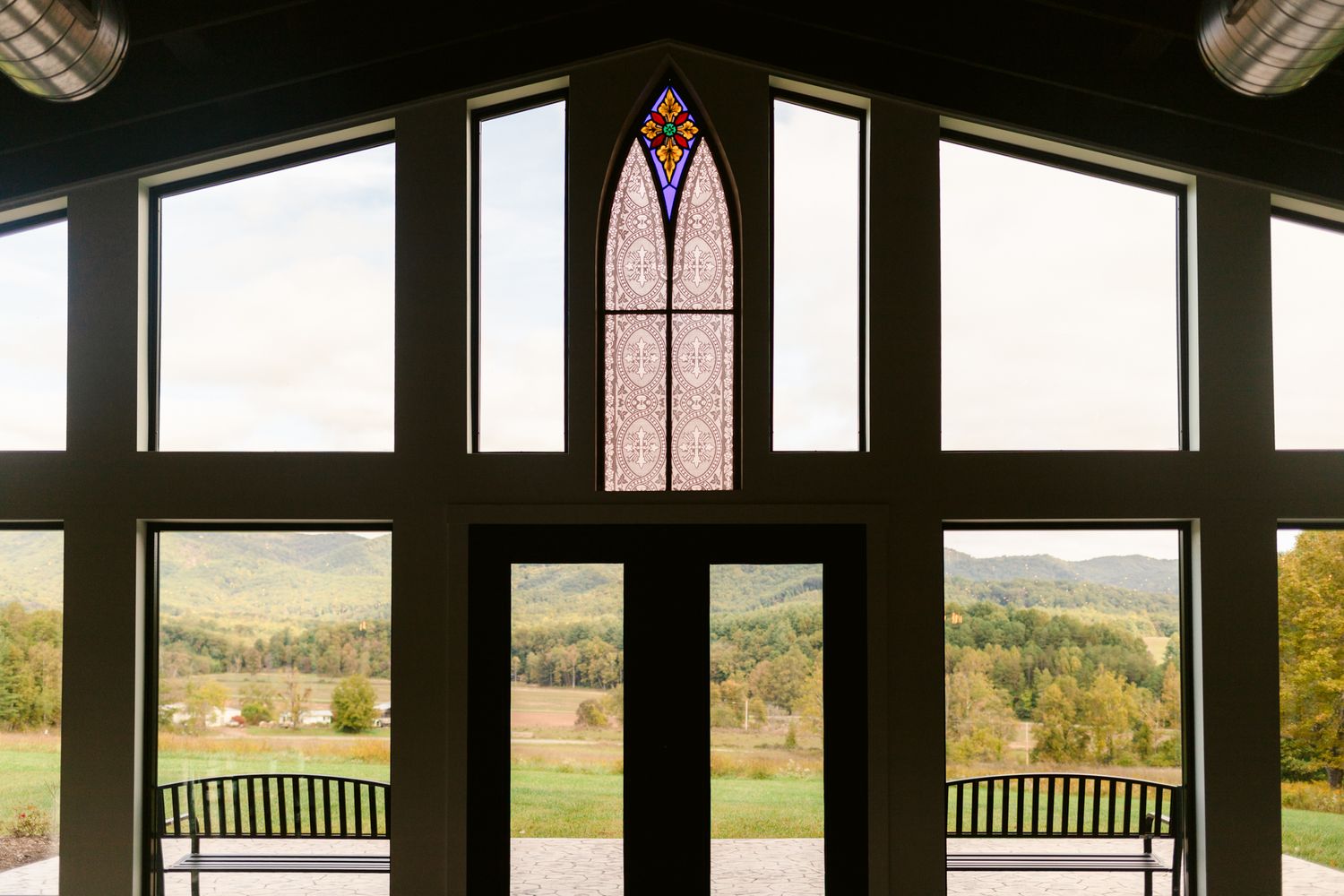 A row of wooden benches faces large windows overlooking a scenic rural landscape with rolling hills and autumn trees.