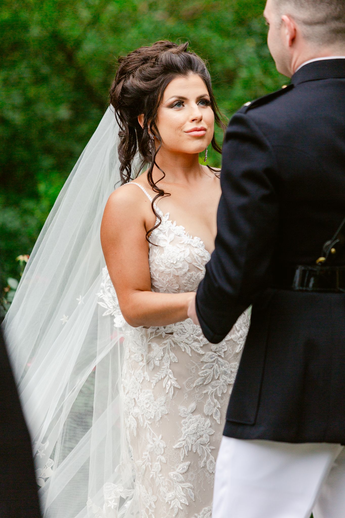 A bride in a detailed lace wedding gown with long veil stands with her groom in military dress during their ceremony.