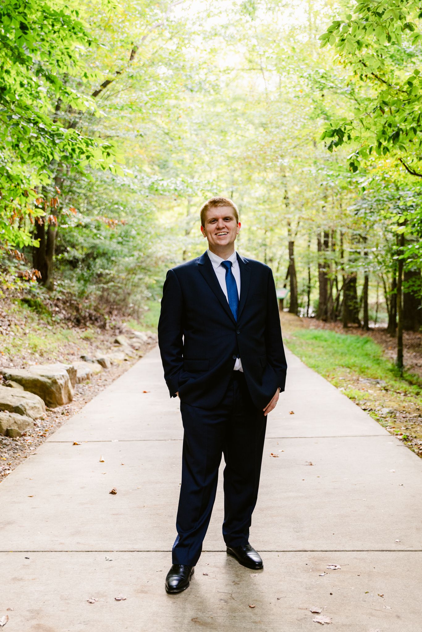 A person in a dark suit stands on a tree-lined path in a park setting during autumn.