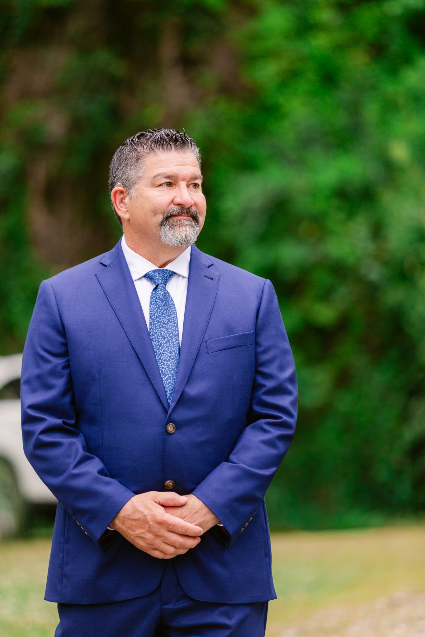 A person wearing a navy blue suit and polka dot tie poses outdoors against a green foliage background.