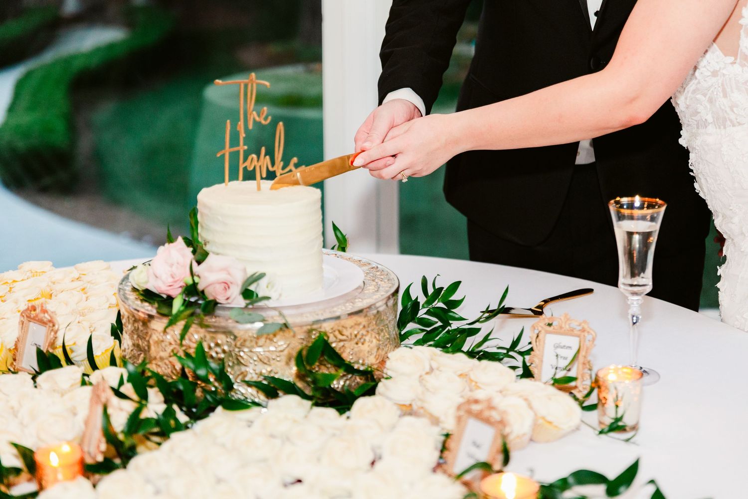 Hands cut into an elegant white wedding cake adorned with fresh flowers and greenery at a formal reception.