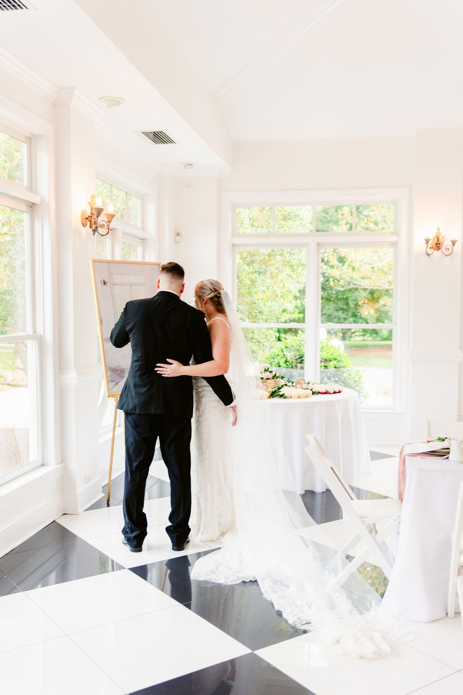 A couple in wedding attire embrace while standing on a black and white checkered floor in a bright sunlit room.