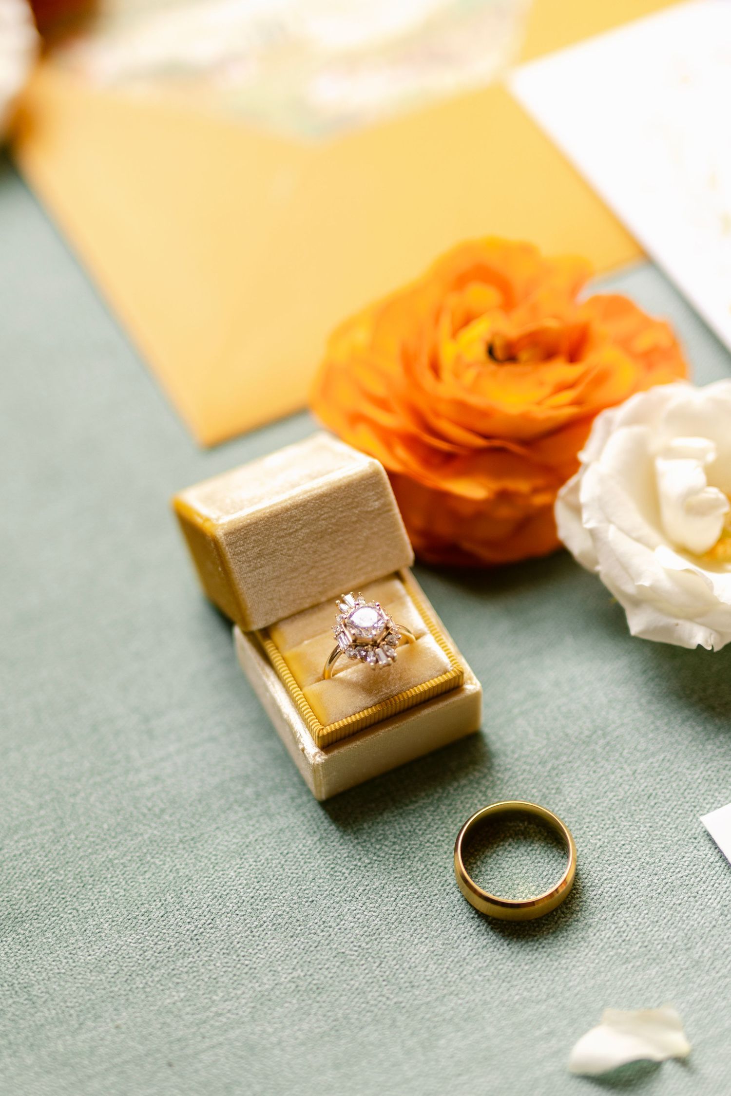 Gold wedding rings and jewelry boxes arranged with orange and white flowers on a sage green textured surface.