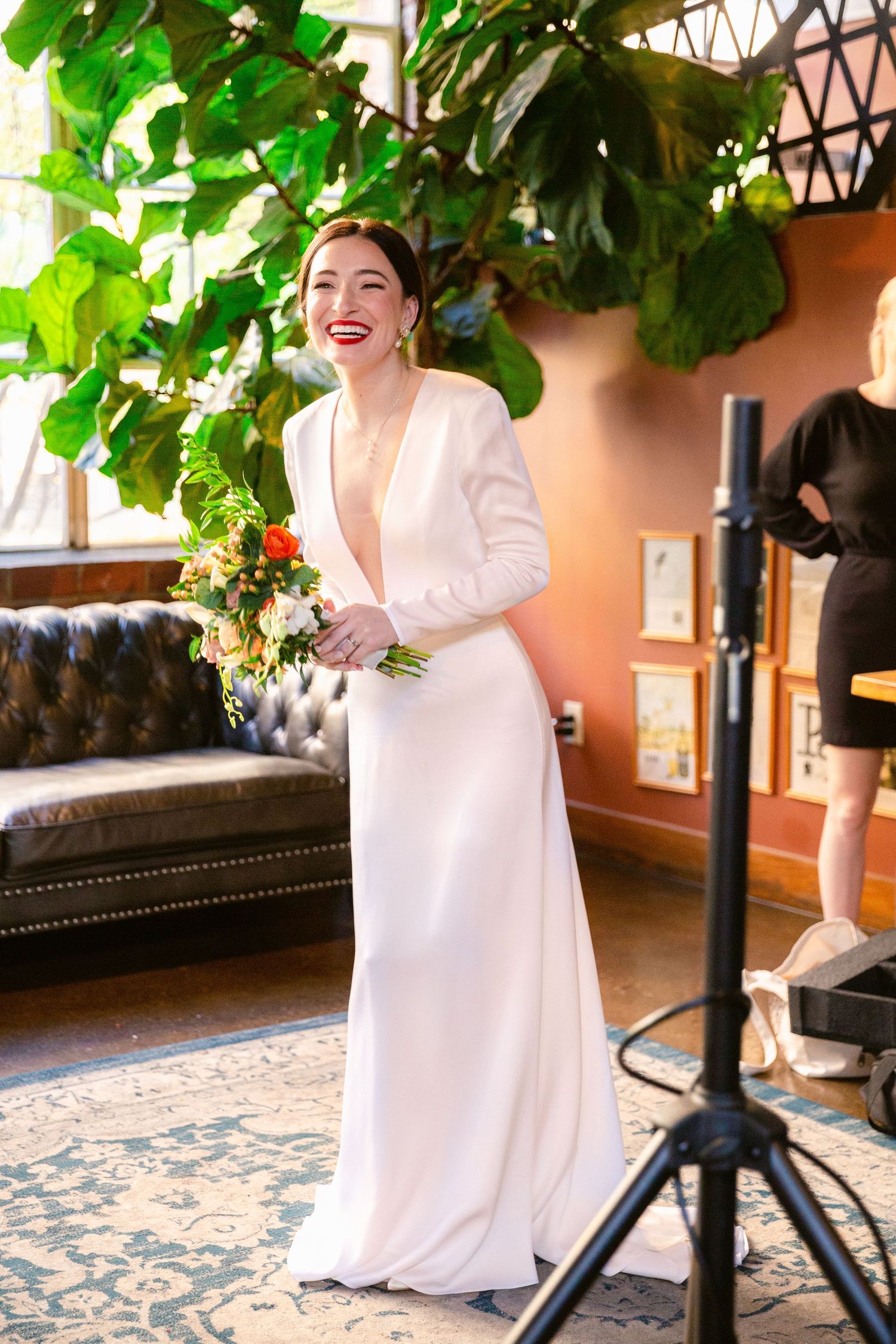 A bride in an elegant long-sleeved white gown laughs joyfully next to a leather couch and lush green plants.