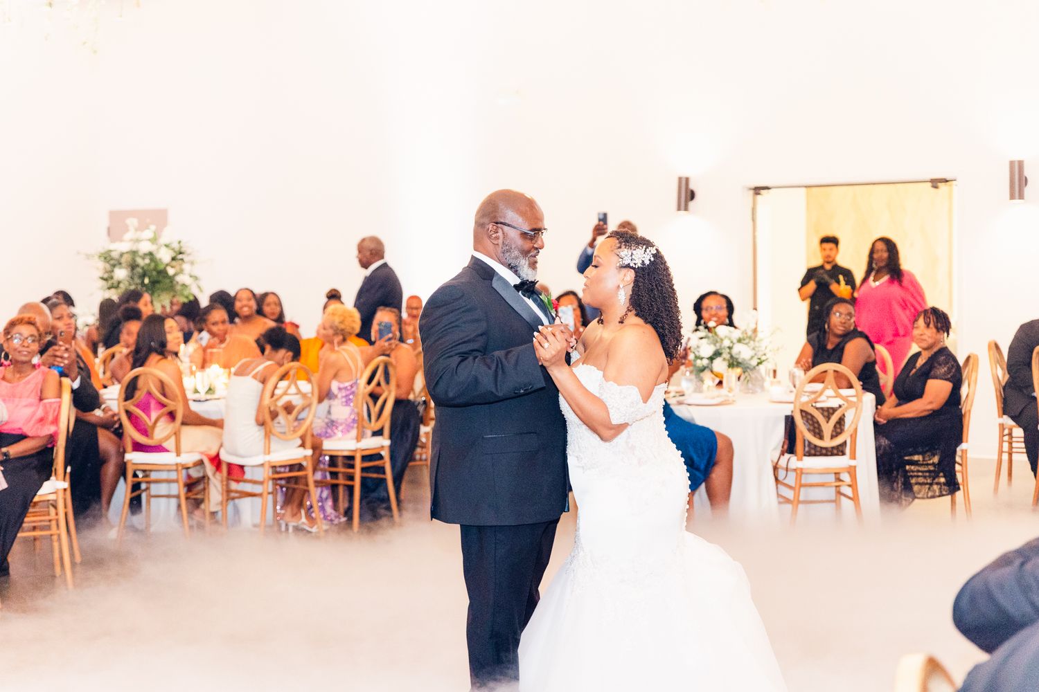 Guests seated in rows at an elegant white wedding venue with bright lighting and floral decorations.