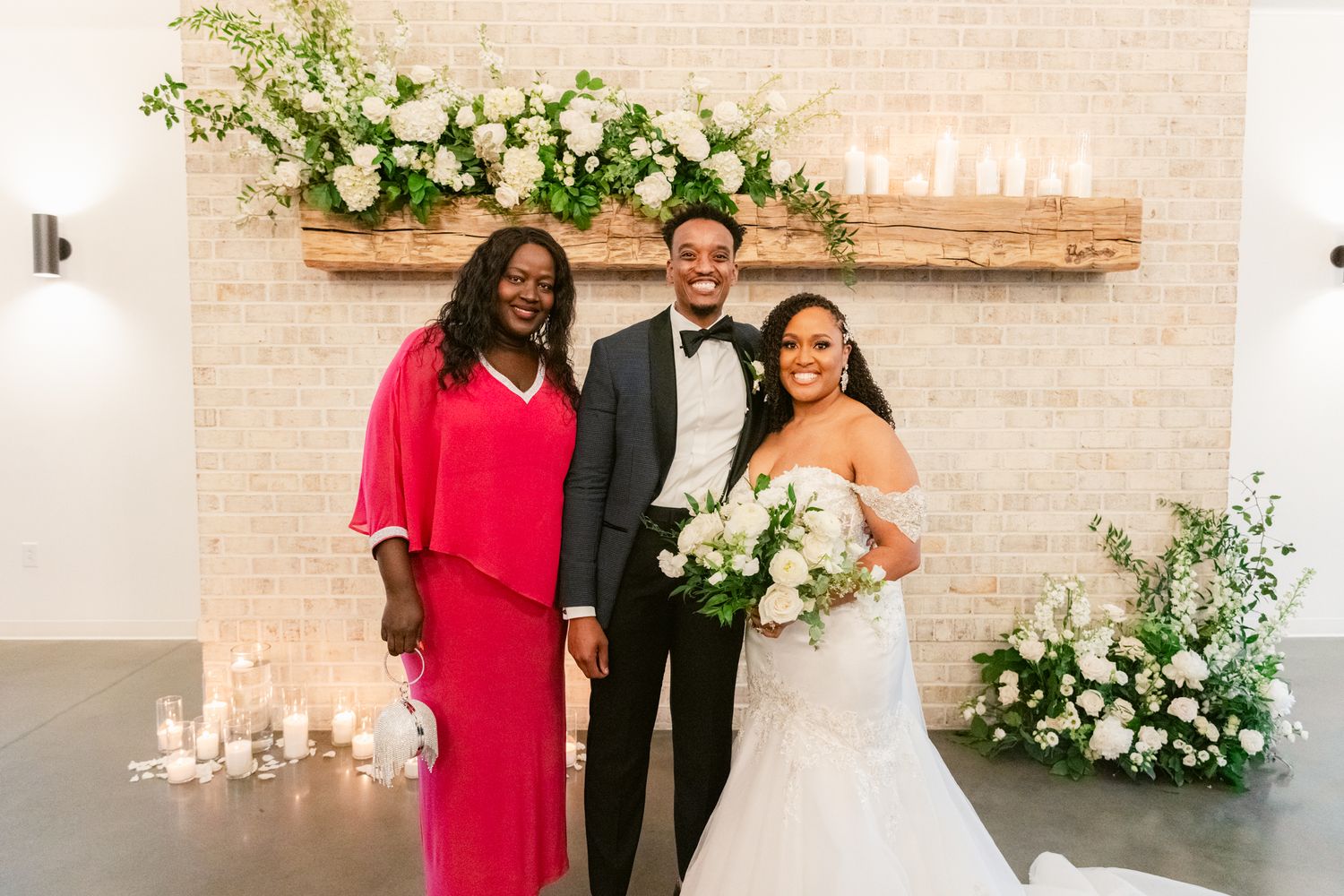 Three people pose together at a wedding celebration with white floral decorations against a light brick wall.