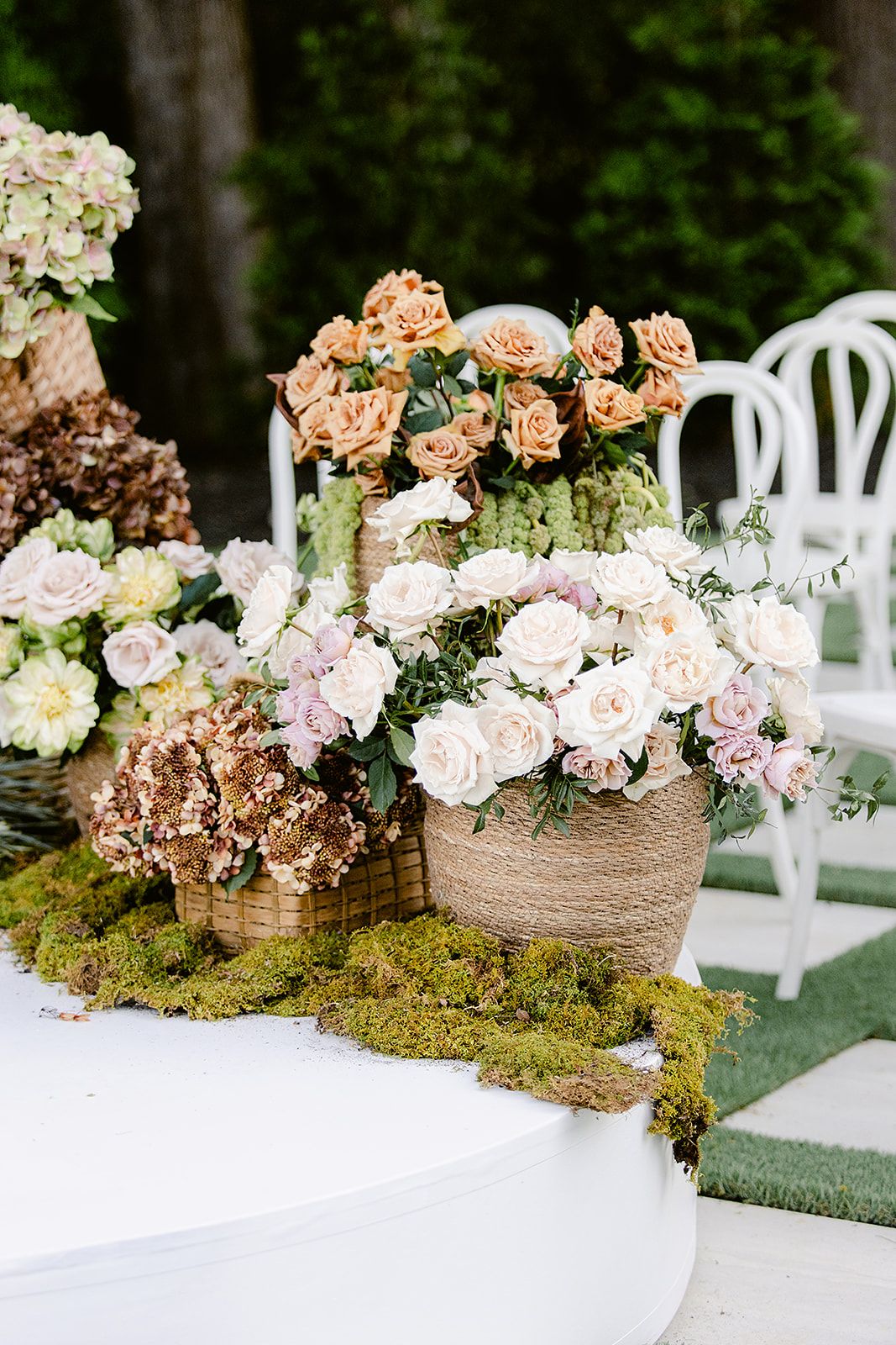 Rustic wedding centerpiece with pink roses and moss arranged in woven baskets on a white surface.