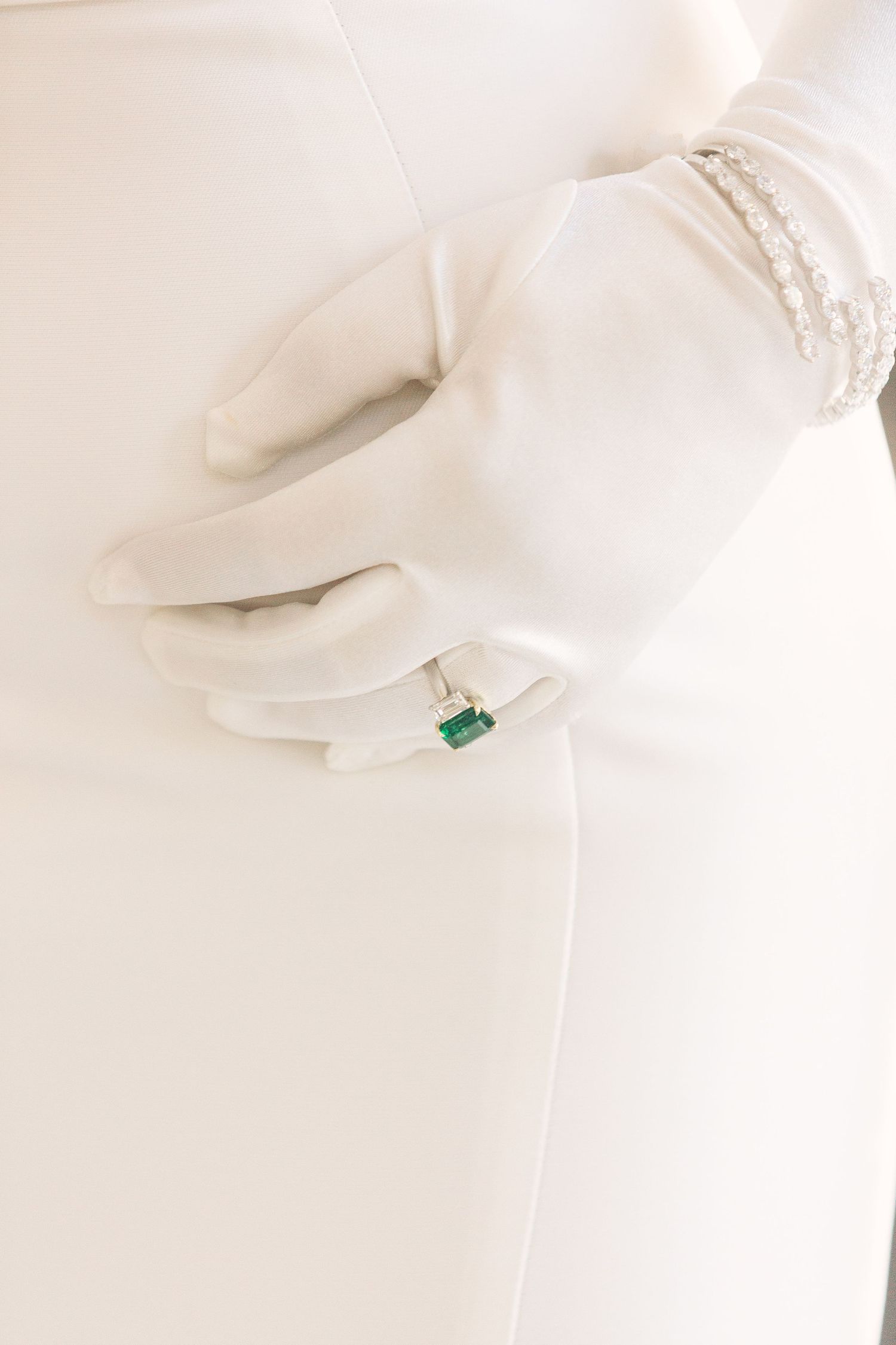 White gloved hands hold a delicate emerald ring against a soft white background.