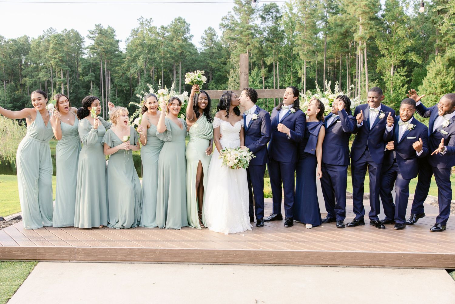 Wedding party members in navy suits and sage green dresses pose together outdoors for formal group photos.
