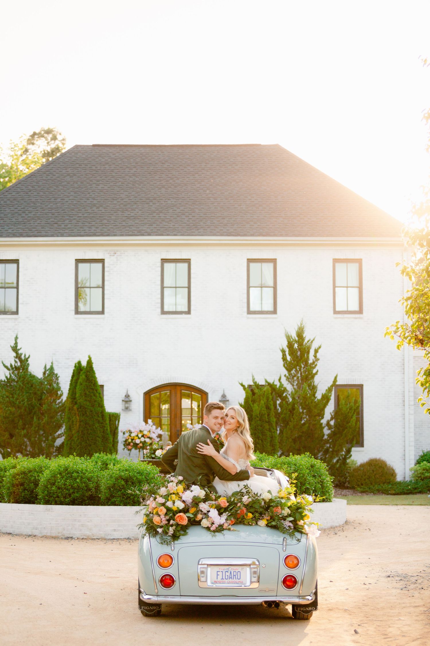 A vintage convertible decorated with floral arrangements is photographed at sunset outside a white wedding venue.