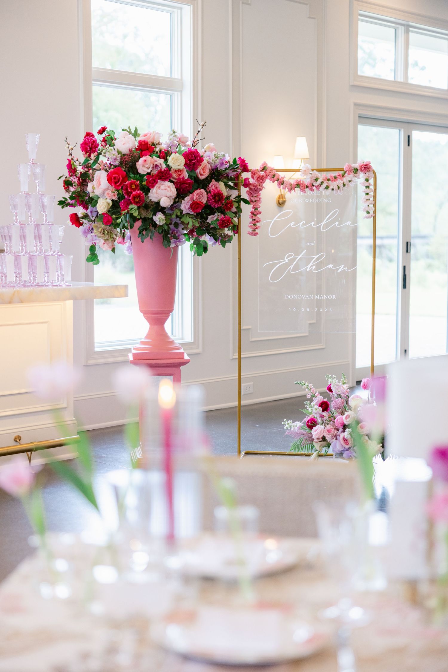 Pink and red floral centerpieces with gold accents decorate an elegant white wedding reception space with natural lighting.