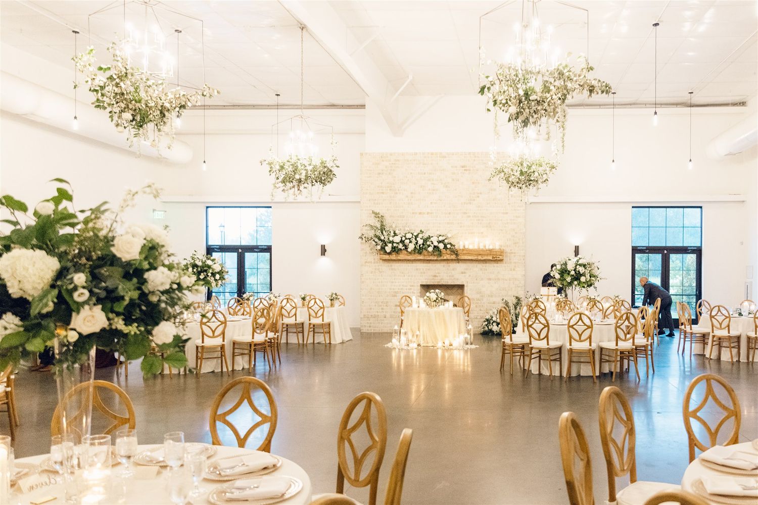 A bright white wedding reception venue with elegant table settings, floral centerpieces, and gold cross-back chairs.
