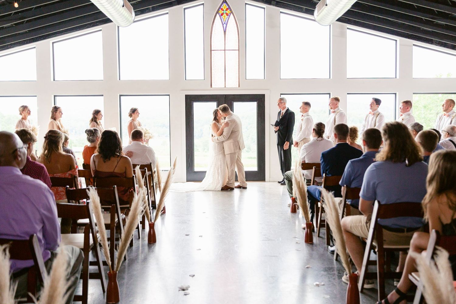 Wedding guests seated in rows at a rustic indoor venue with large windows and natural light.