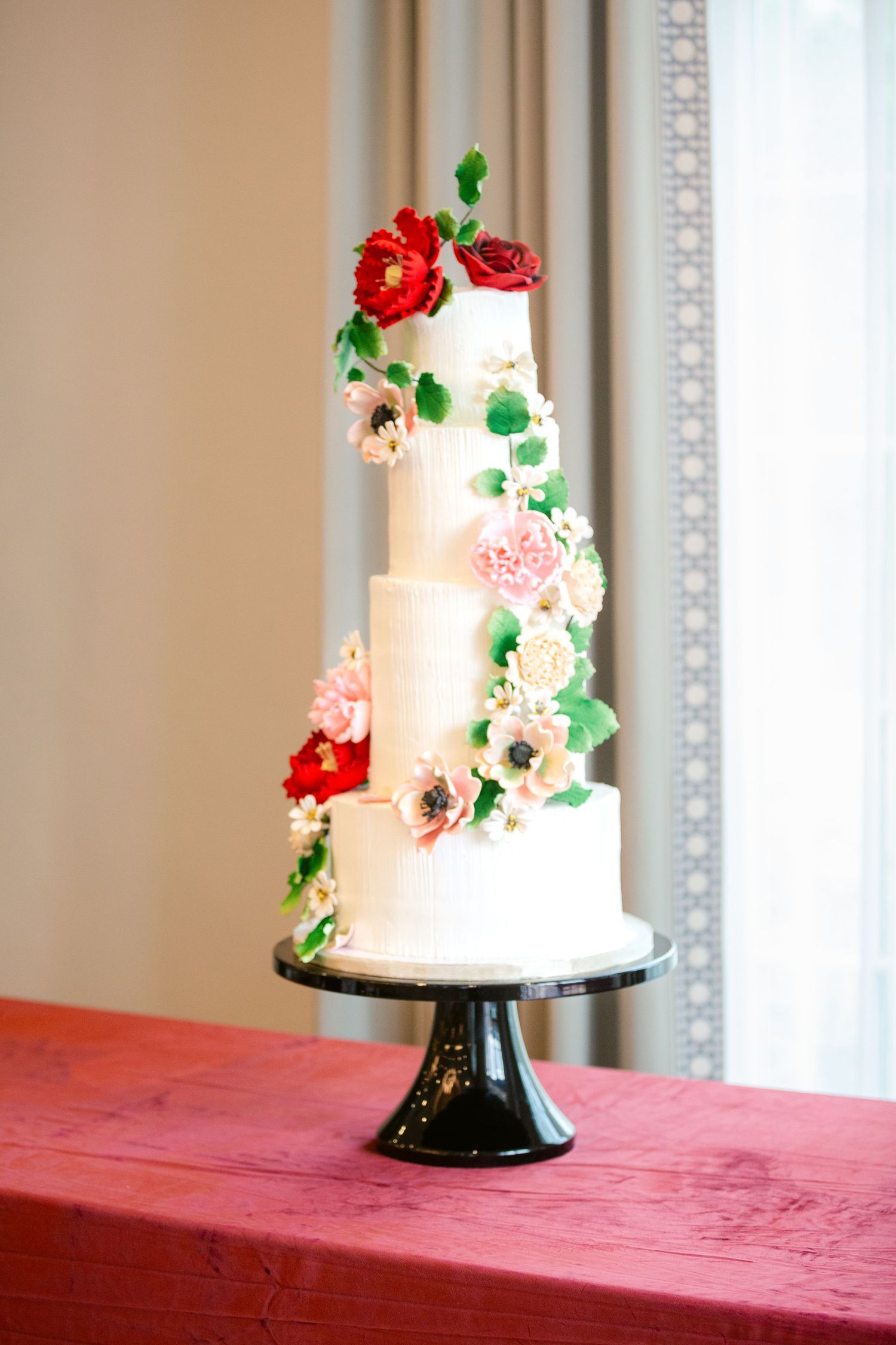 Elegant white wedding cake decorated with red and pink sugar flowers displayed on a black pedestal against a light background.