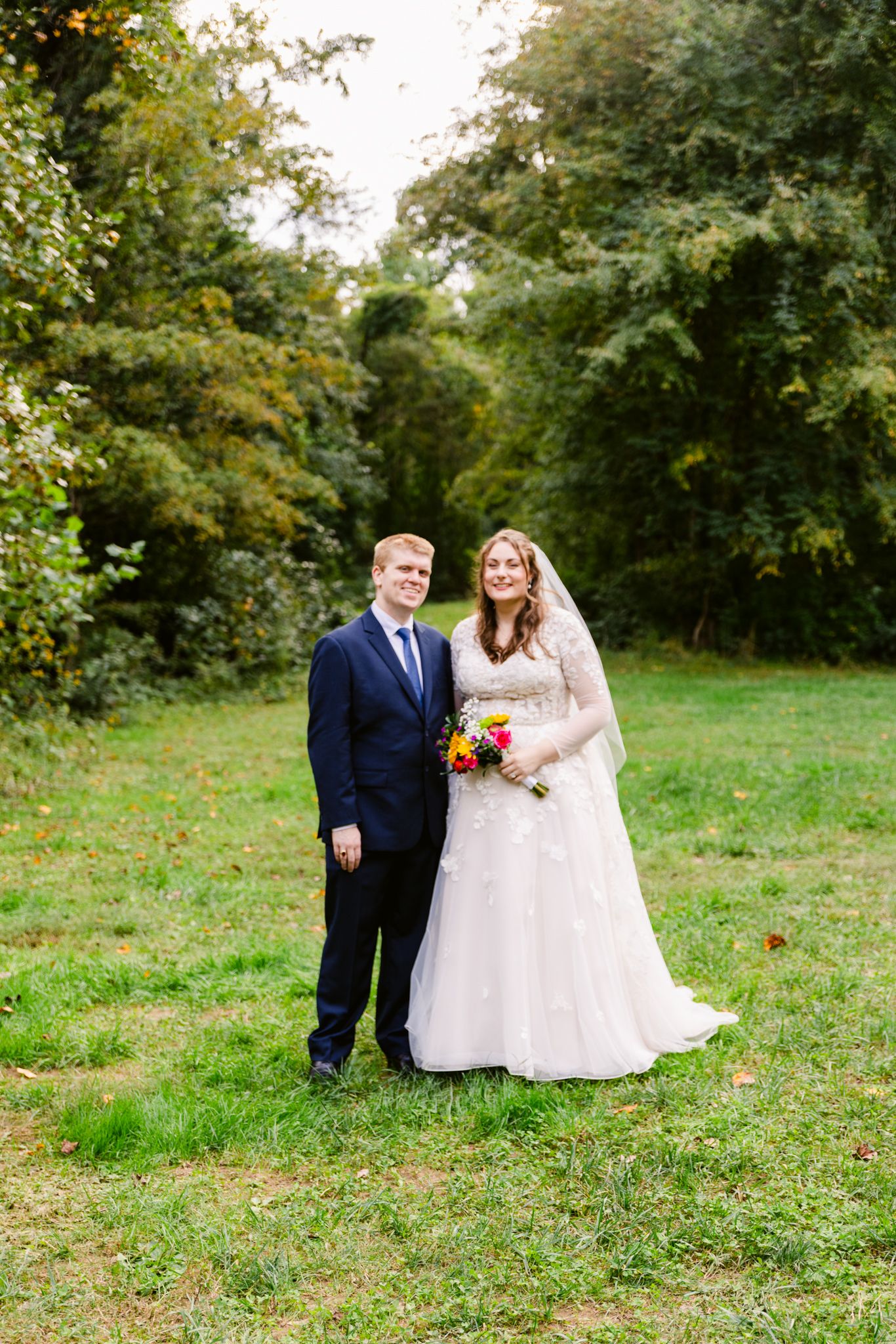 A couple in wedding attire poses together on lush green grass surrounded by autumn trees in the background.