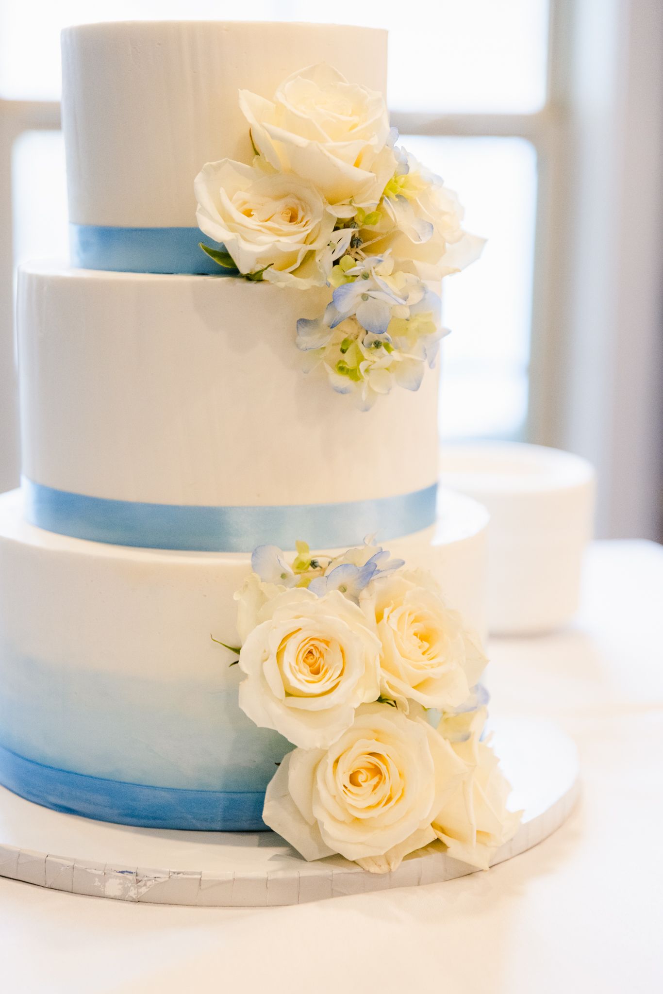 Elegant three-tier white wedding cake decorated with light blue ribbons and cream-colored roses on a marble base.